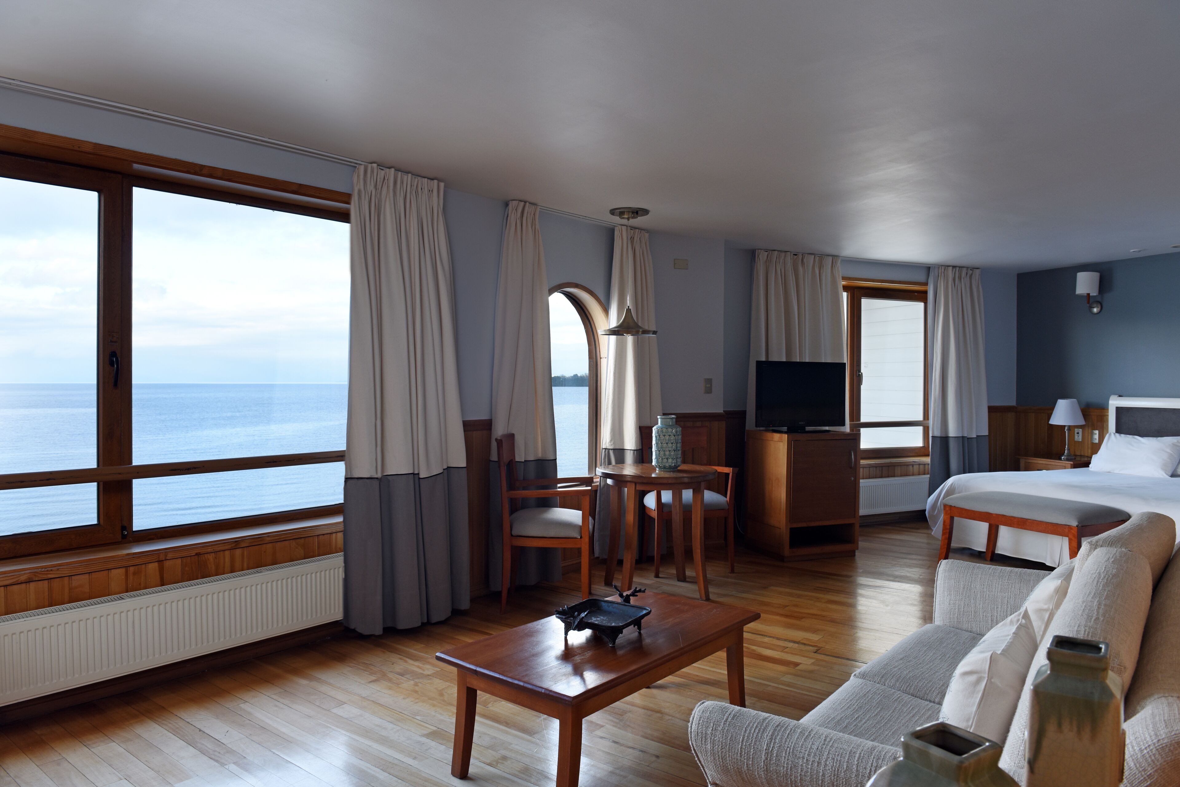 A hotel room with a large window, wooden furniture, a bed, a desk, and a television.