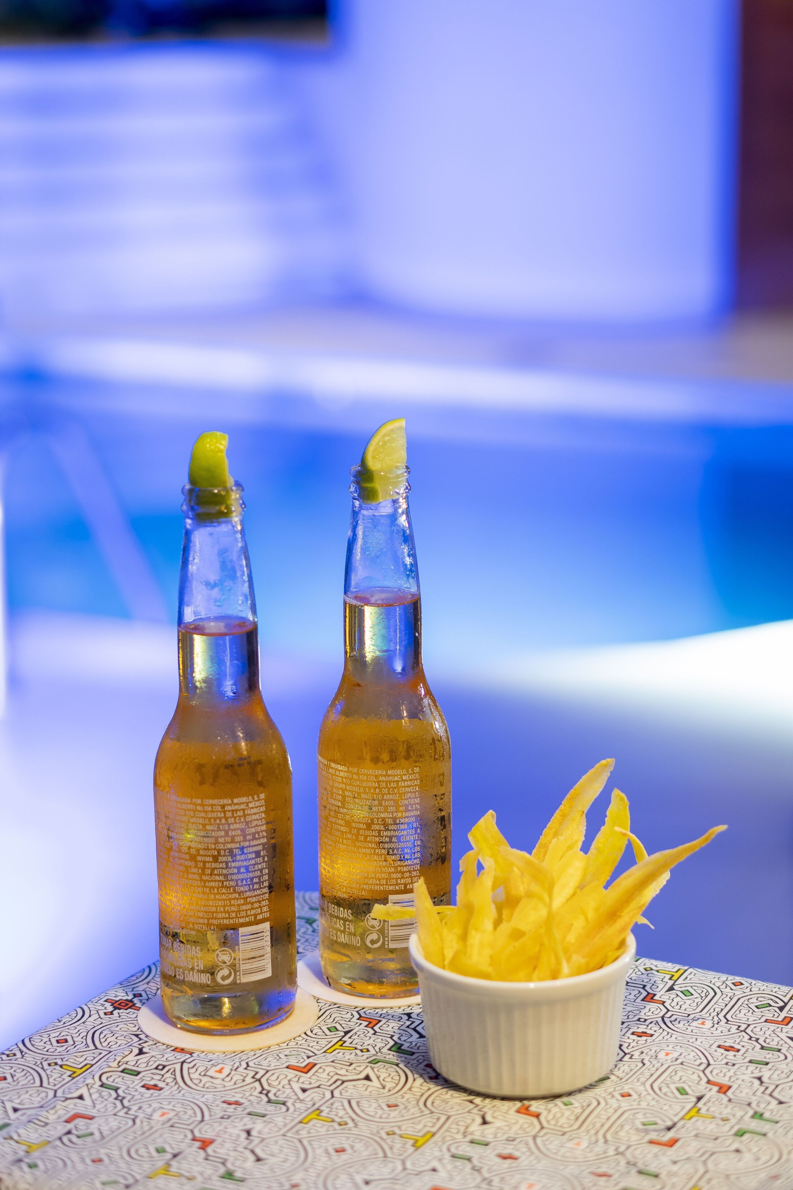 Two bottles of a beverage with a lemon garnish, a bowl of lemon slices, and a patterned tablecloth.