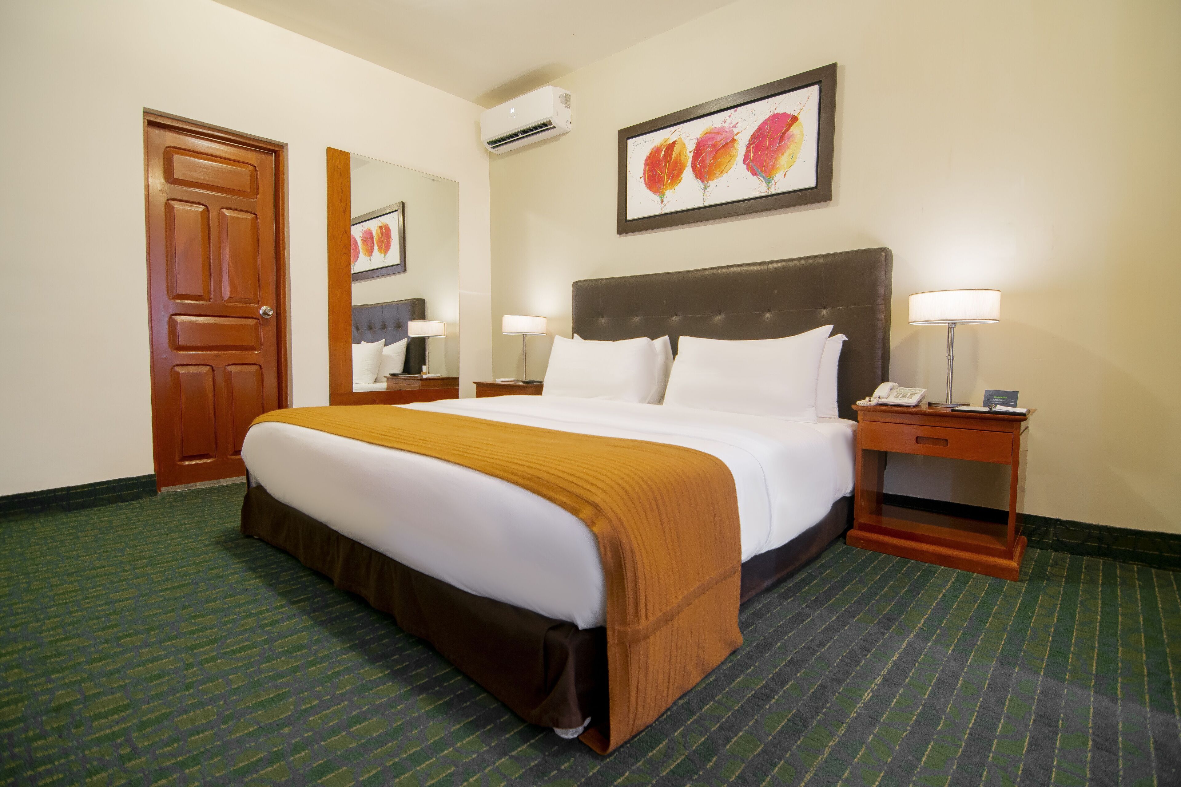 A hotel room with a large bed, a nightstand, a bedside table, a wall-mounted air conditioner, and a framed floral artwork above the bed.