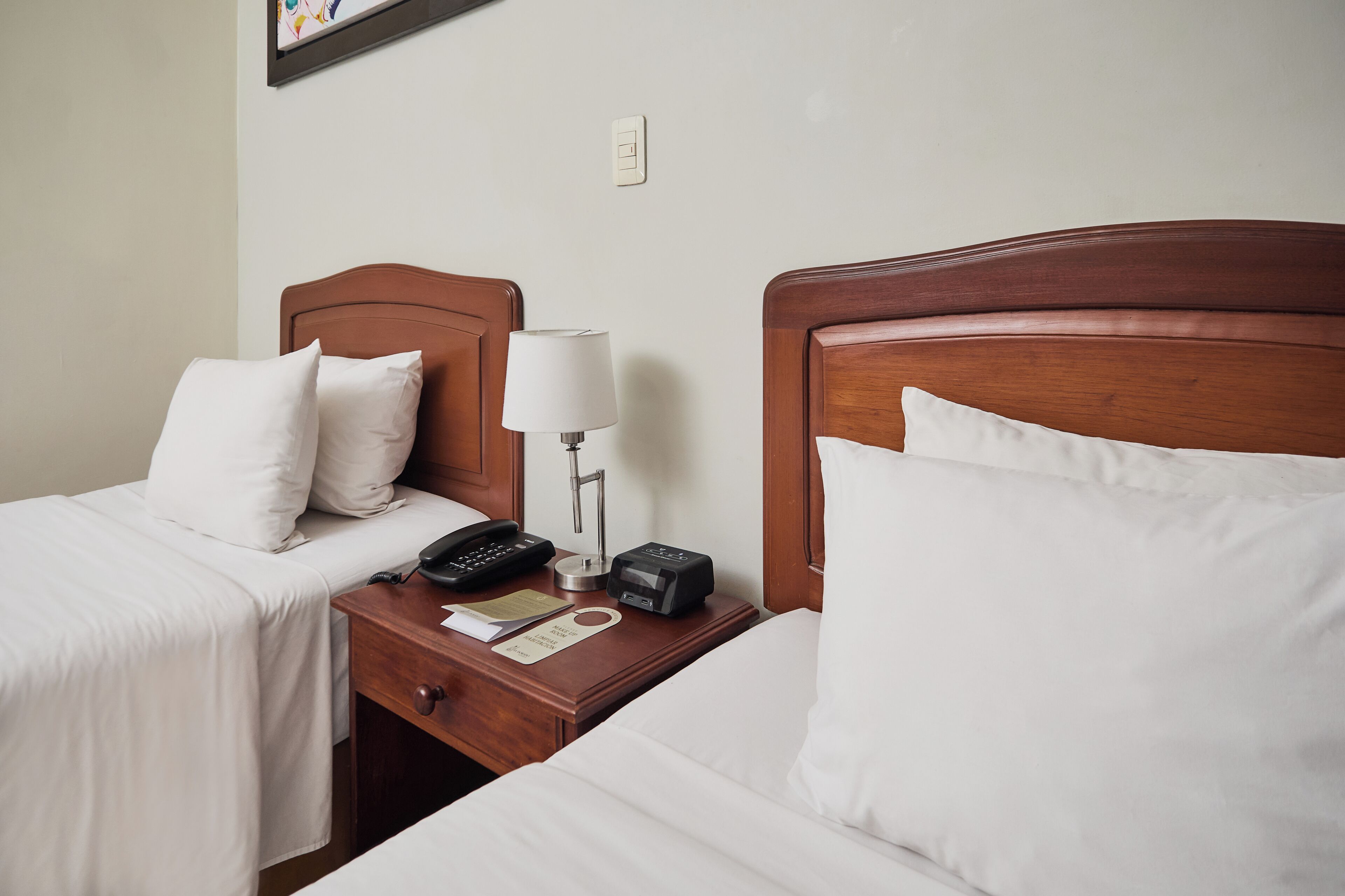 A hotel room with two single beds, a wooden headboard, a bedside table with a lamp, a phone, and a notepad.