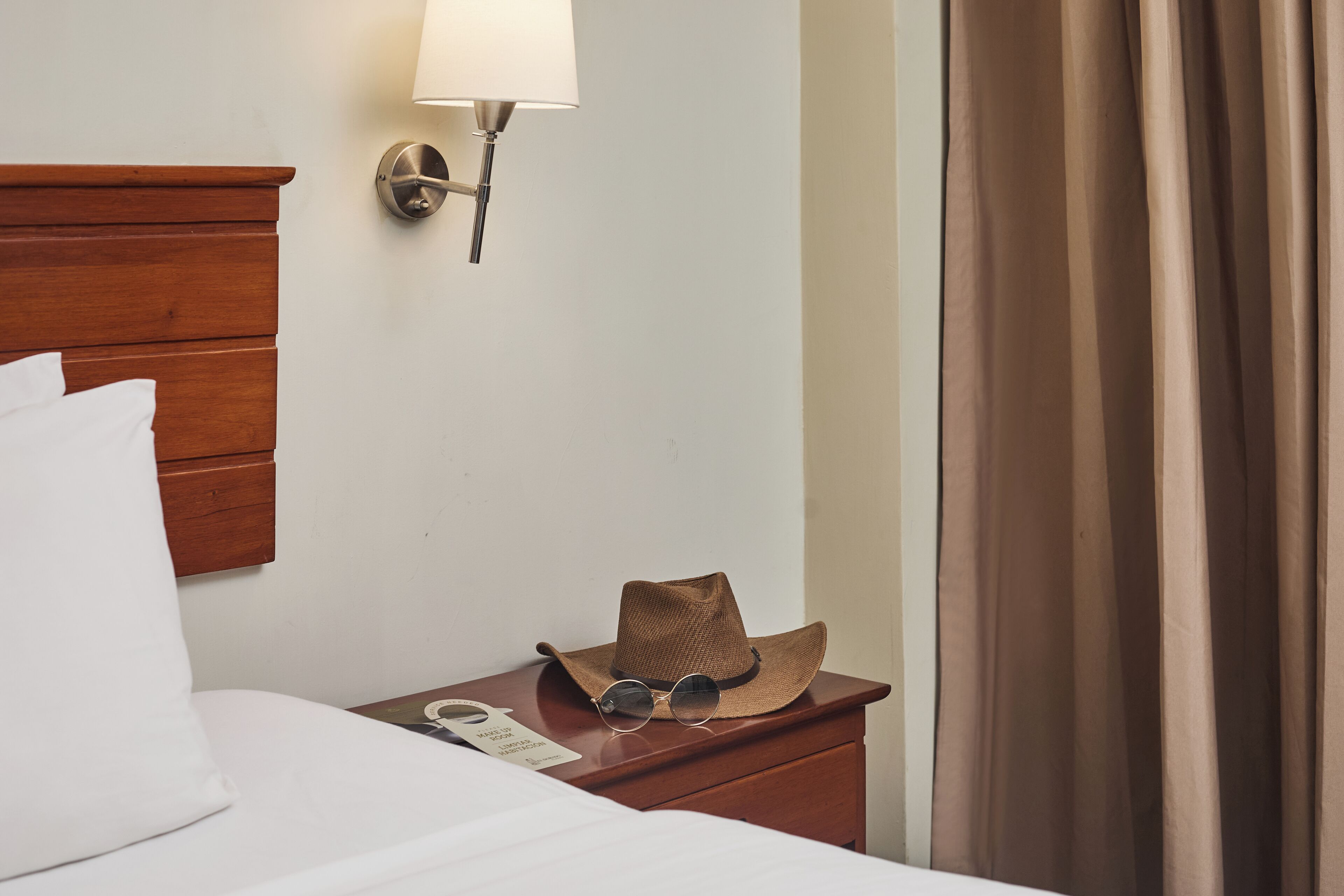 A neatly made bed with white linens, a wooden nightstand with a hat and sunglasses, and a wall-mounted lamp.