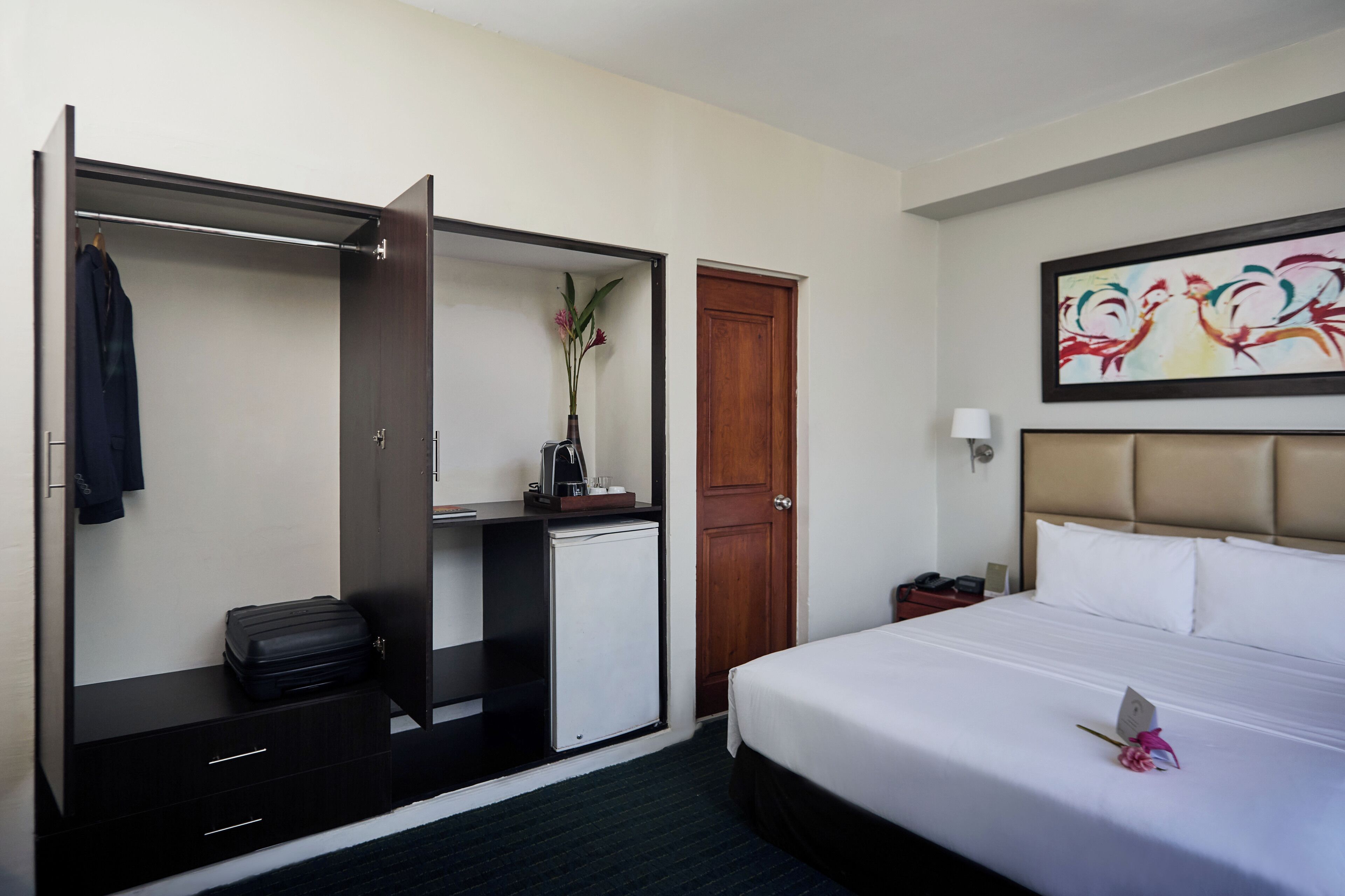 A hotel room with a bed, a wardrobe, a desk, a small refrigerator, and a painting on the wall.