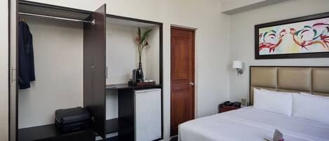 A hotel room with a bed, a wardrobe, a desk, a small refrigerator, and a painting on the wall.