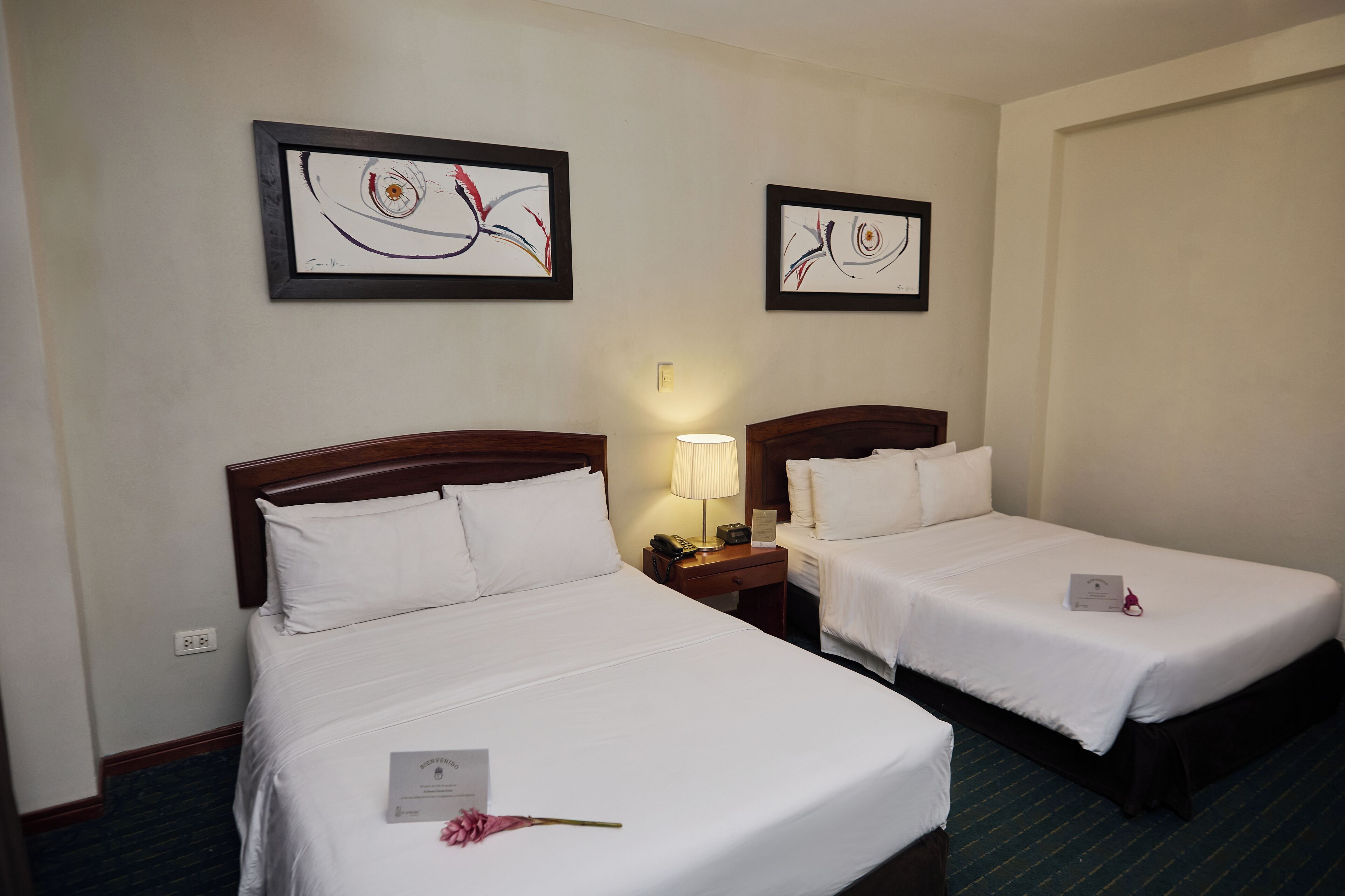 A hotel room with two single beds, a bedside table with a lamp, and two framed abstract paintings on the wall.