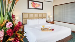 A hotel room with a large bed, a headboard, a bedside table with a lamp, a framed artwork on the wall, and a window with a view of the sky.