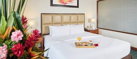 A hotel room with a large bed, a headboard, a bedside table with a lamp, a framed artwork on the wall, and a window with a view of the sky.