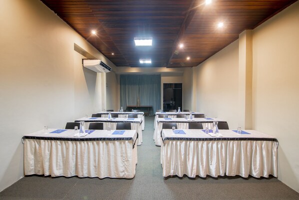 Un salón de conferencias con seis mesas, cada una con mantel blanco y posavasos azules. El salón tiene techo de madera y un ventanal grande con cortinas.