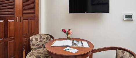 A room with a wooden wardrobe, a round wooden table with two patterned chairs, a flat-screen TV mounted on the wall, and a thermostat on the wall.