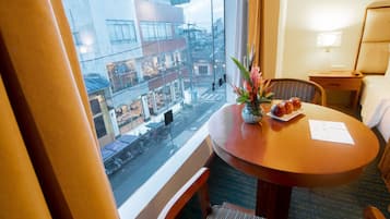 A hotel room with a large window, a round wooden table with a vase of flowers, a chair, and a small bedside table with a lamp.
