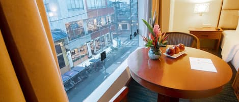Habitación de hotel con un ventanal grande, una mesa redonda de madera con un florero, una silla y una mesita de noche con una lámpara.