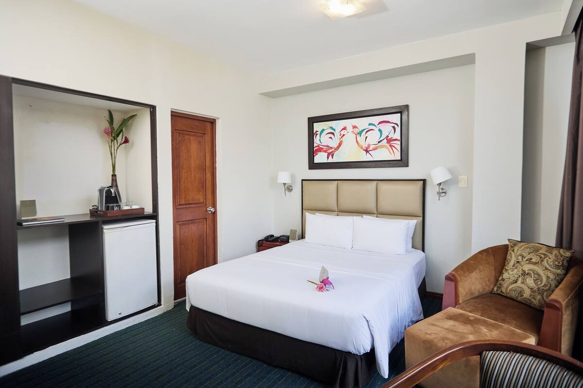 Ein Hotelzimmer mit Bett, Sessel, Schreibtisch mit Lampe, Telefon und einem Bild an der Wand.