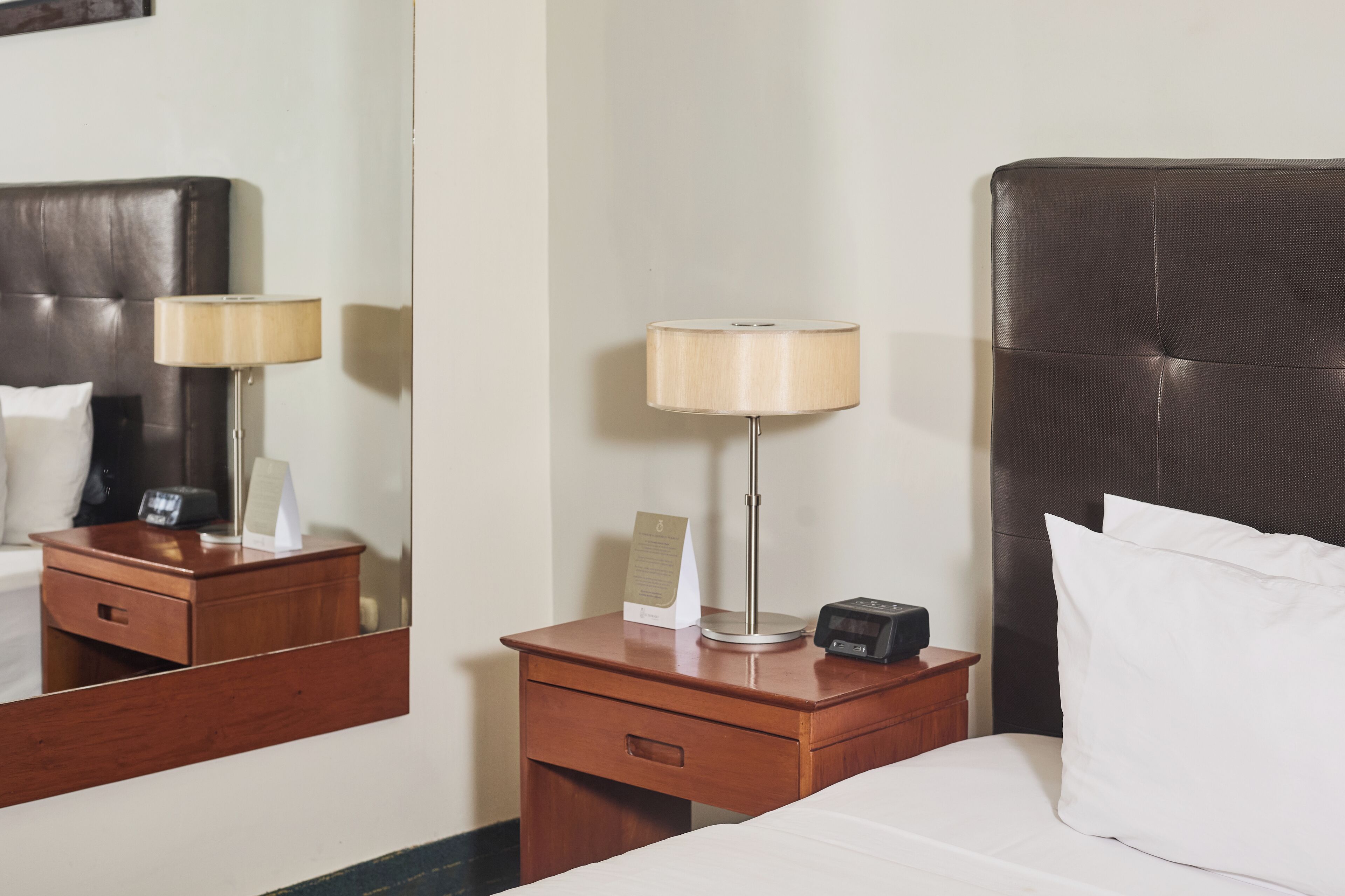 A hotel room with a bed, a nightstand with a lamp, a clock, and a mirror.