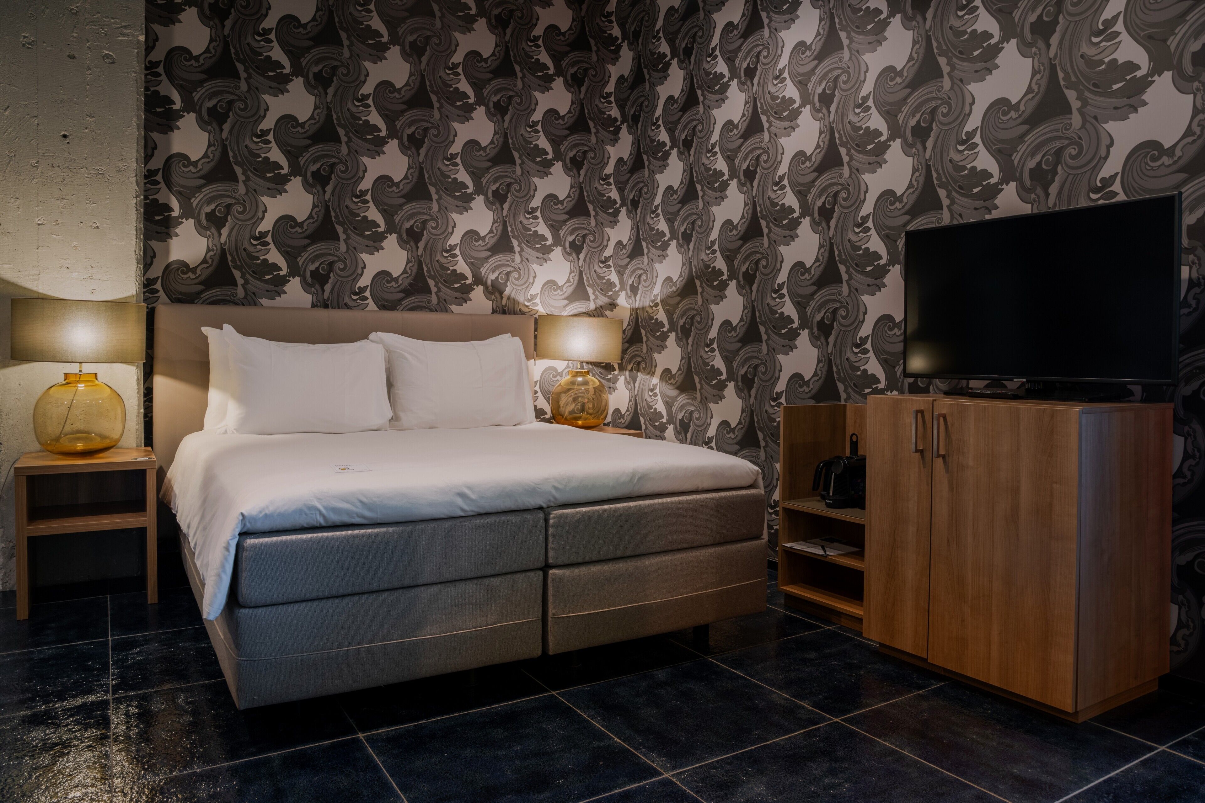 Une chambre à coucher avec un lit, deux lampes de chevet, un téléviseur, une armoire en bois et un papier peint à motifs.