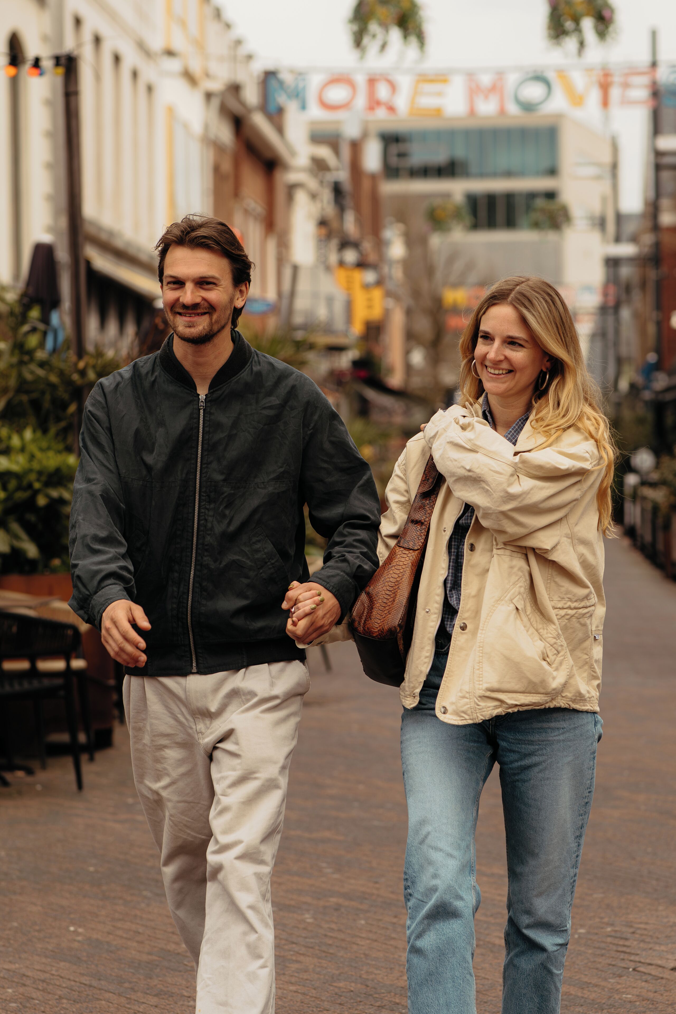 Um casal caminhando em uma rua de paralelepípedos, com edifícios e uma placa que diz 'MORE MOVIE'.