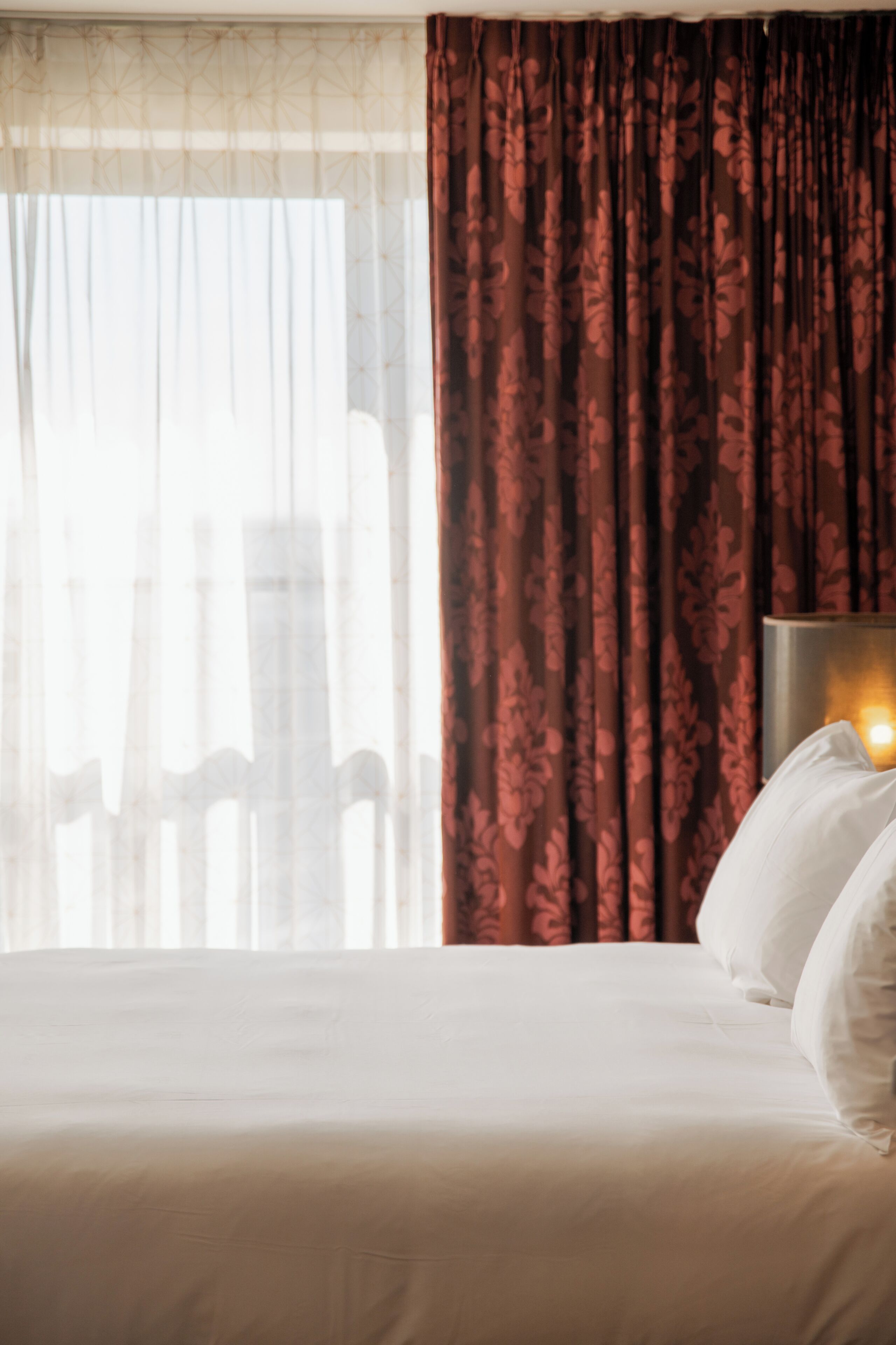 Hotelkamer met een bed, nachtkastje en raam met dunne gordijnen en een patroonkleurig gordijn.