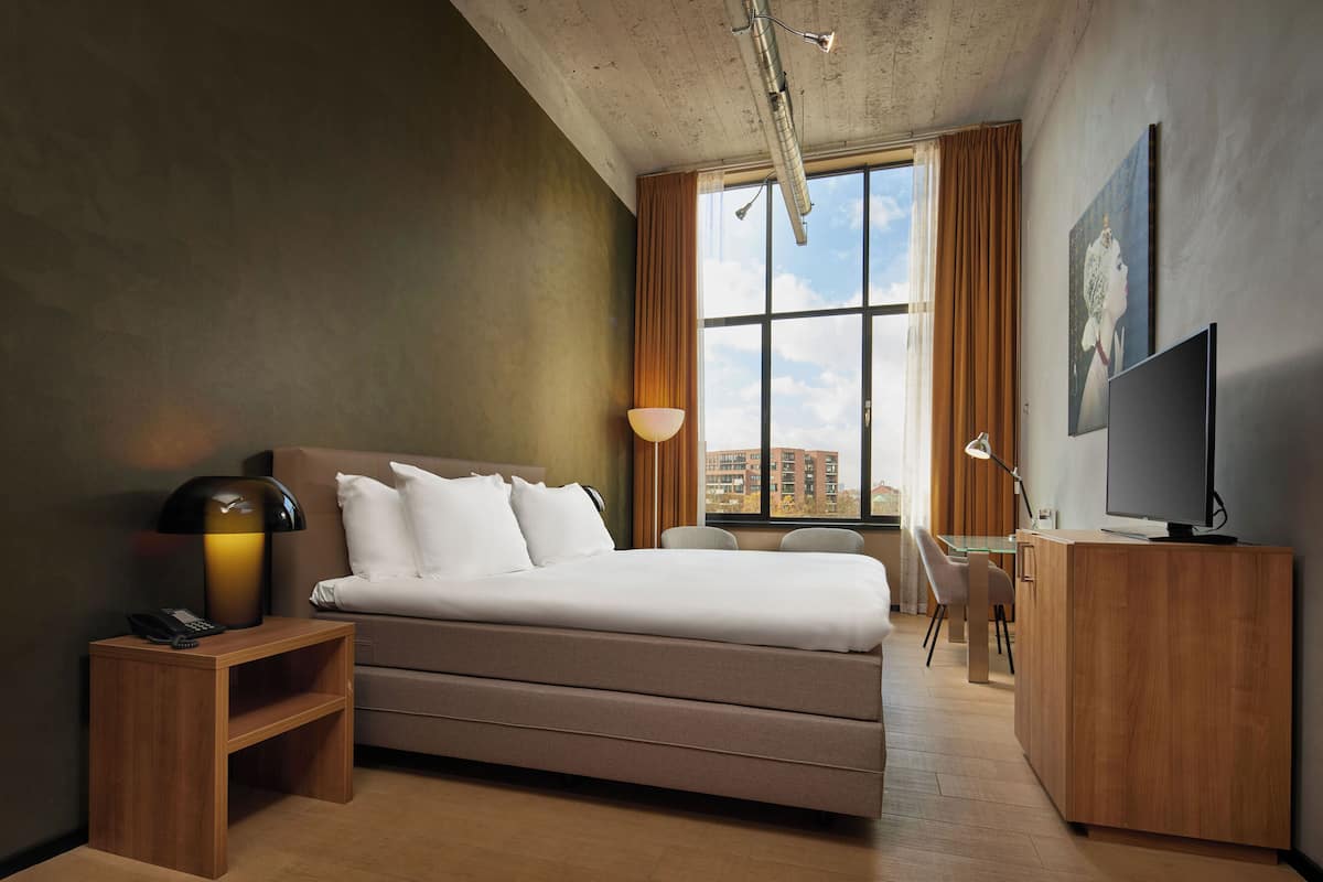 A modern hotel room with a large bed, a desk, a television, and a window with curtains.