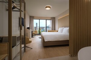 Quarto de hotel moderno com uma cama grande, uma escrivaninha com cadeira, um guarda-roupa e vista da cidade pela janela.