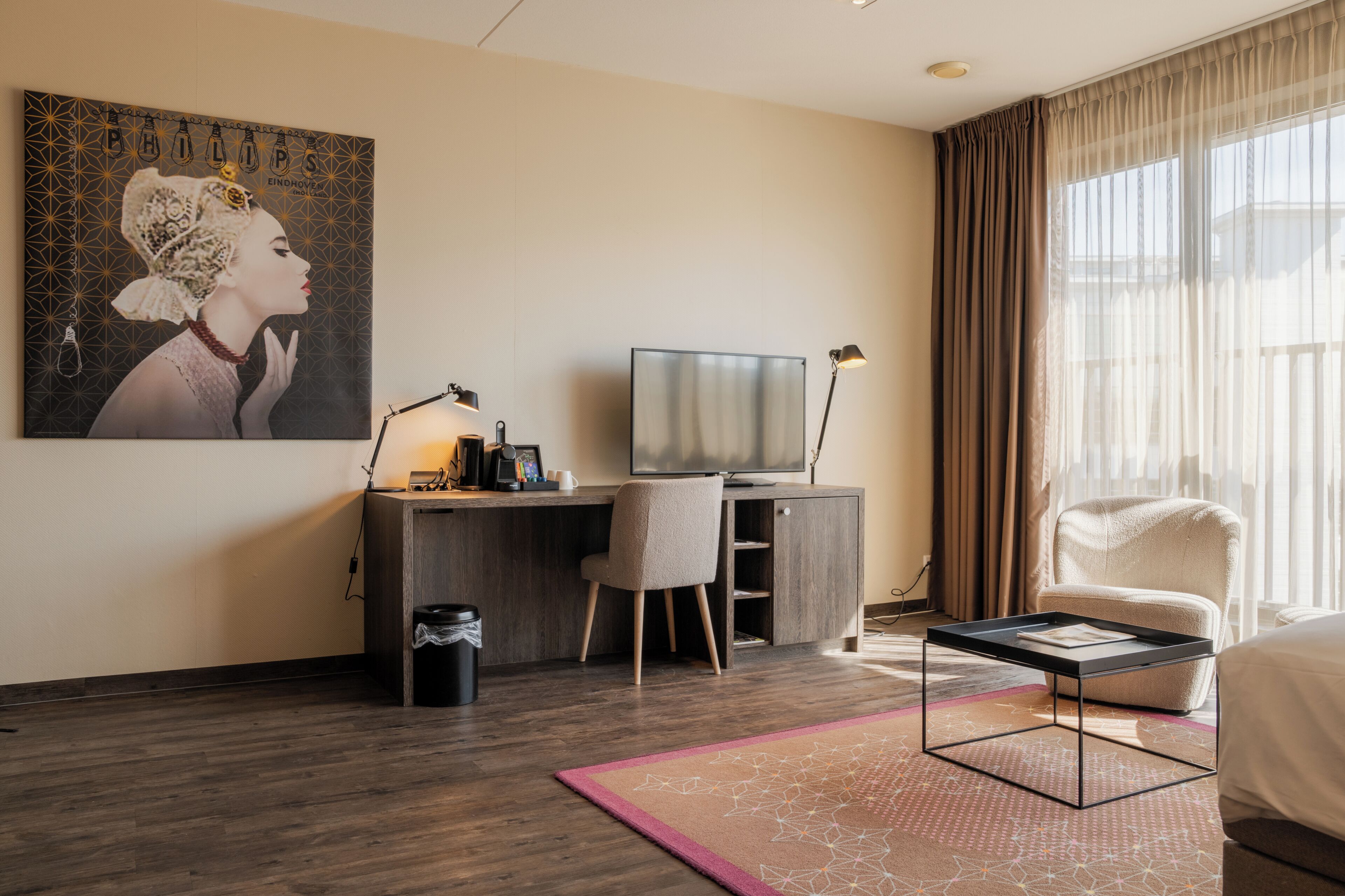 Habitación de hotel con escritorio, silla, televisor y una ventana grande con cortinas.