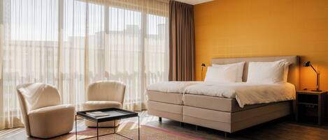 A modern hotel room with a large bed, two armchairs, a small table, and a nightstand with a lamp.