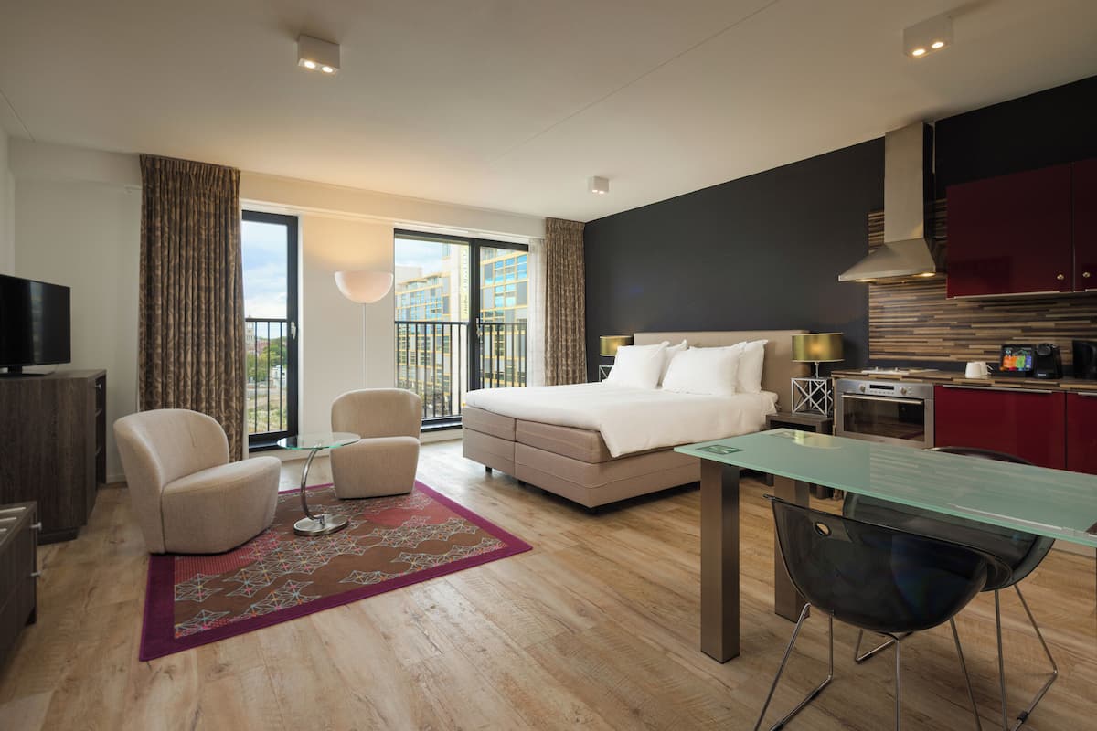 A modern hotel room with a bed, seating area, kitchenette, and a balcony with a city view.