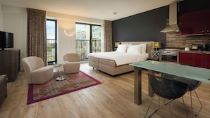 A modern hotel room with a bed, seating area, kitchenette, and a balcony with a city view.