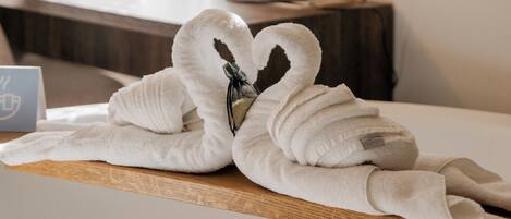 A neatly made bed with a towel arranged in a swan shape, a bedside table with a lamp, and a view of the window with curtains.