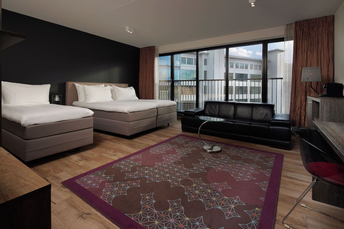 A modern hotel room with two beds, a sofa, a desk, and a large window with a view of buildings.
