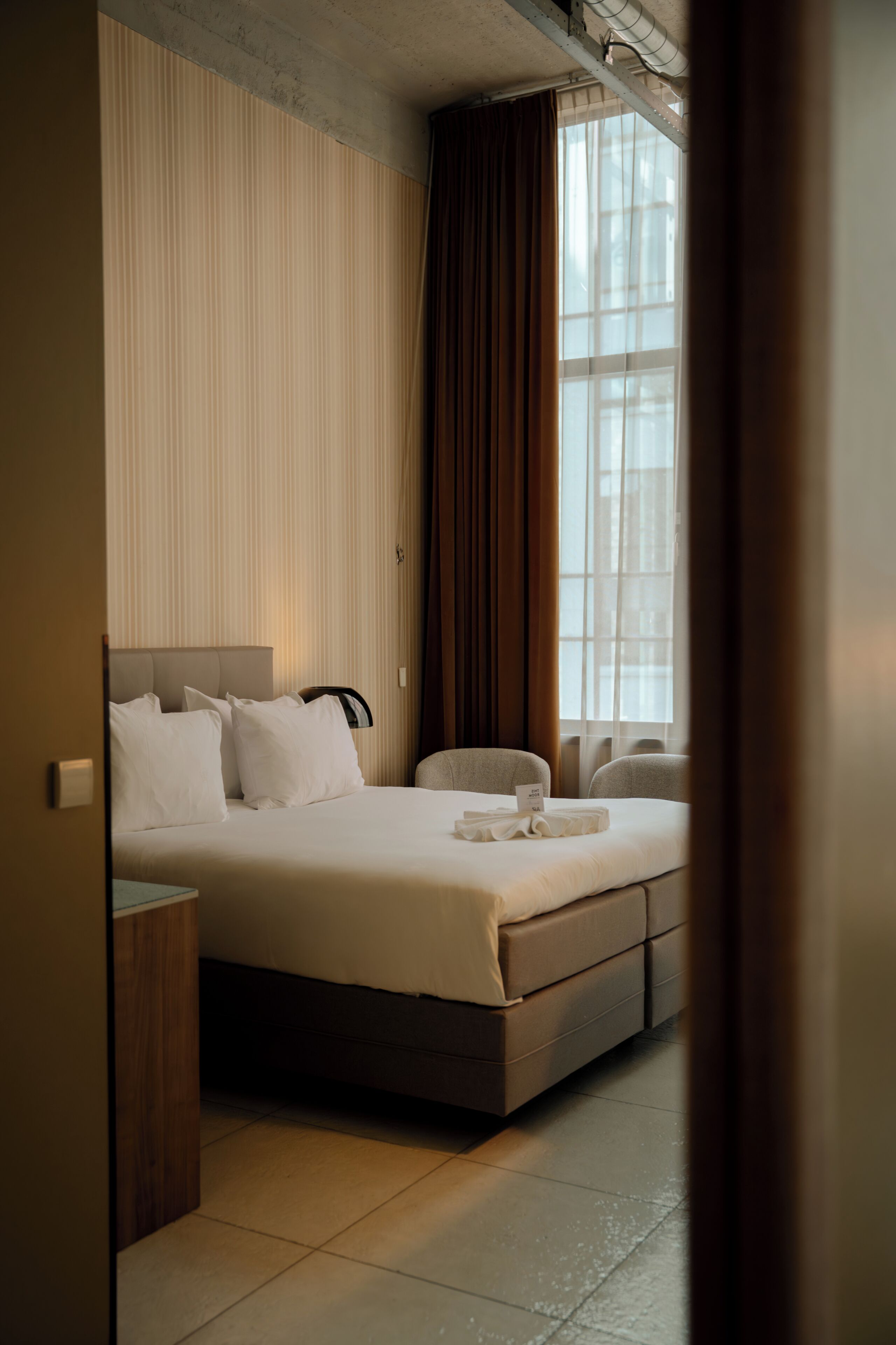 A hotel room with a bed, bedside table, and a window with curtains.
