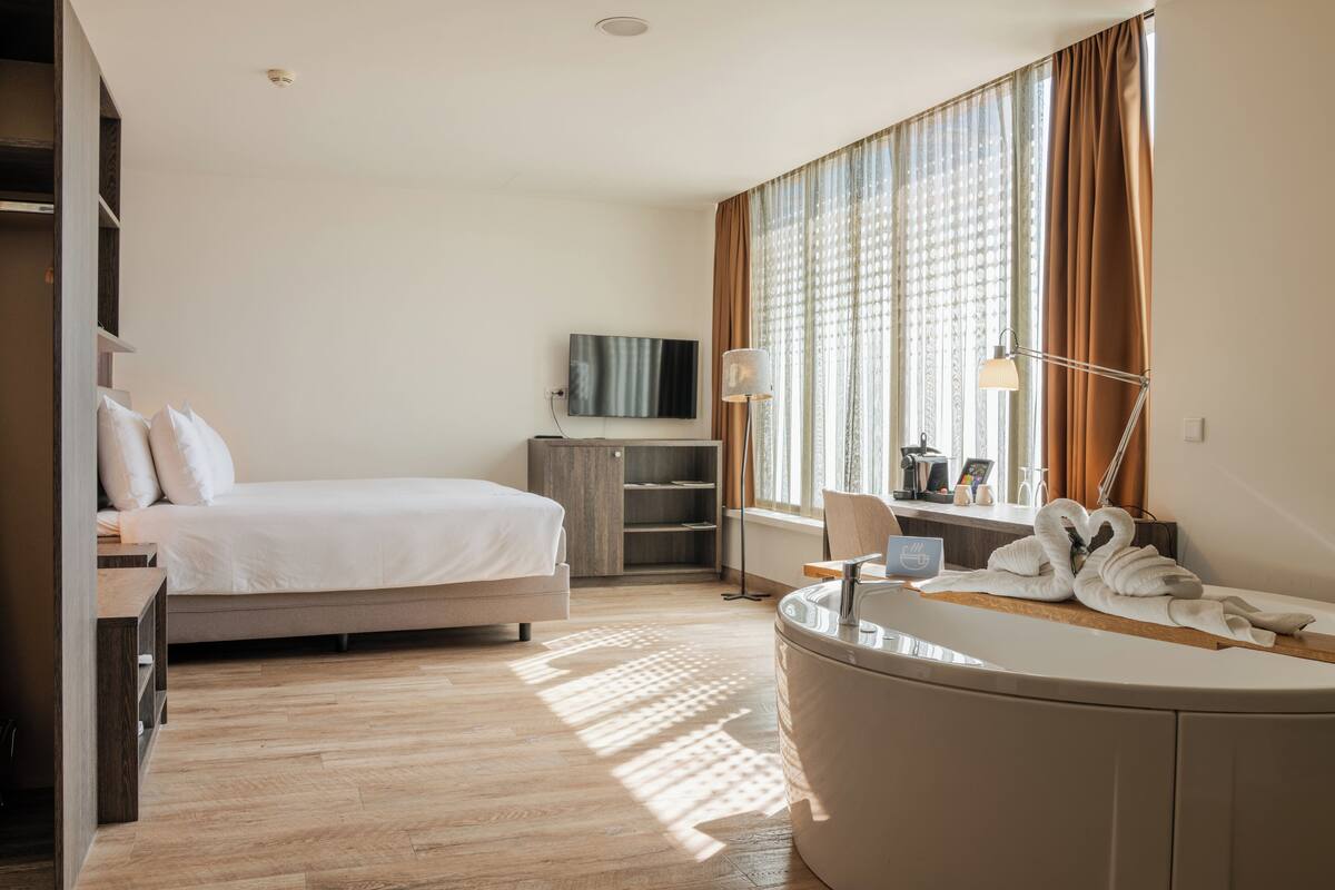 A hotel room with a bed, a vanity with a mirror, a bathtub, a television, and a window with curtains.