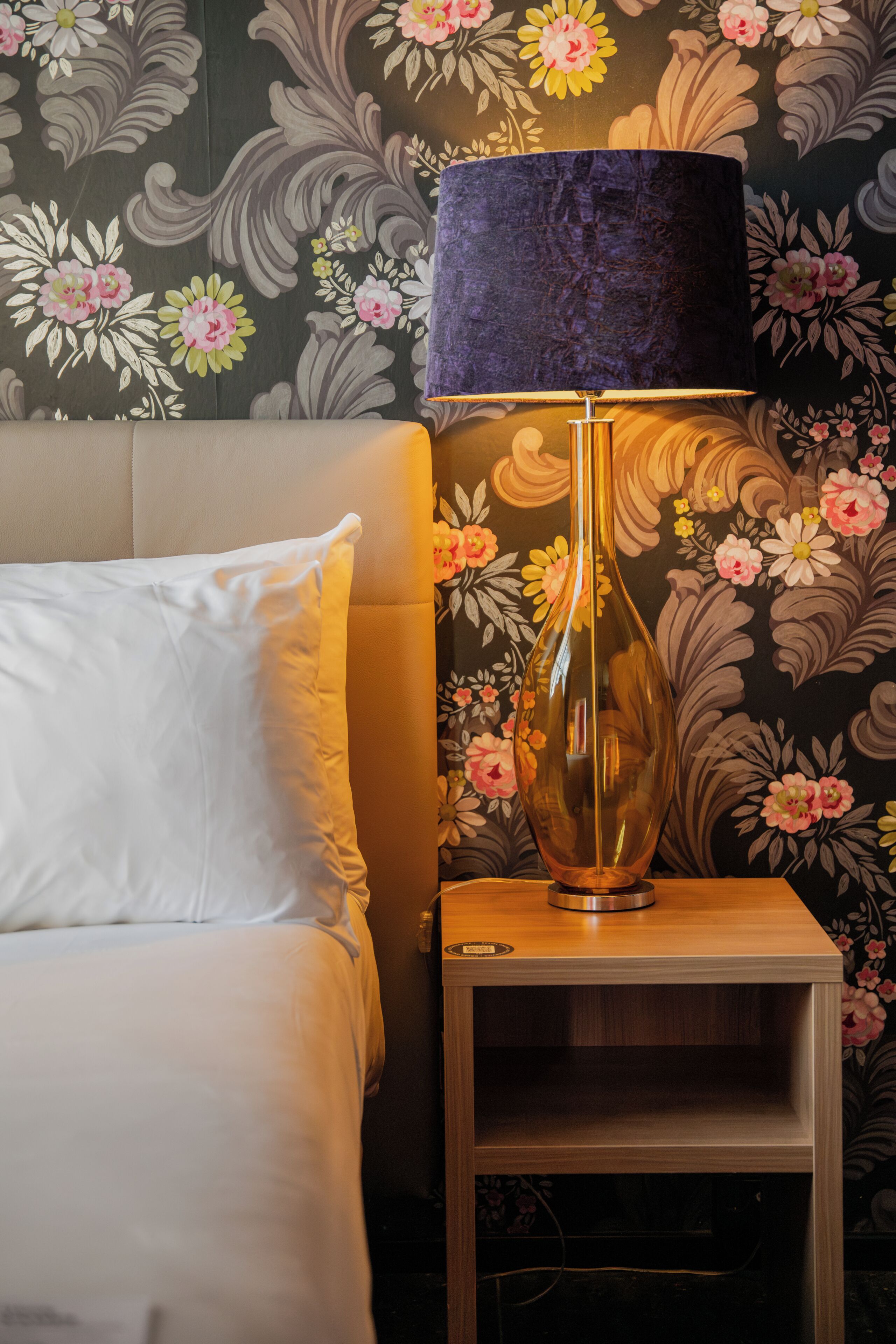 A neatly made bed with white linens, a dark blue lampshade, and a wooden nightstand with a vase.