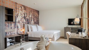 A modern hotel room with a large bathtub, a bed with white linens, a television, and a wall with a geometric patterned artwork.