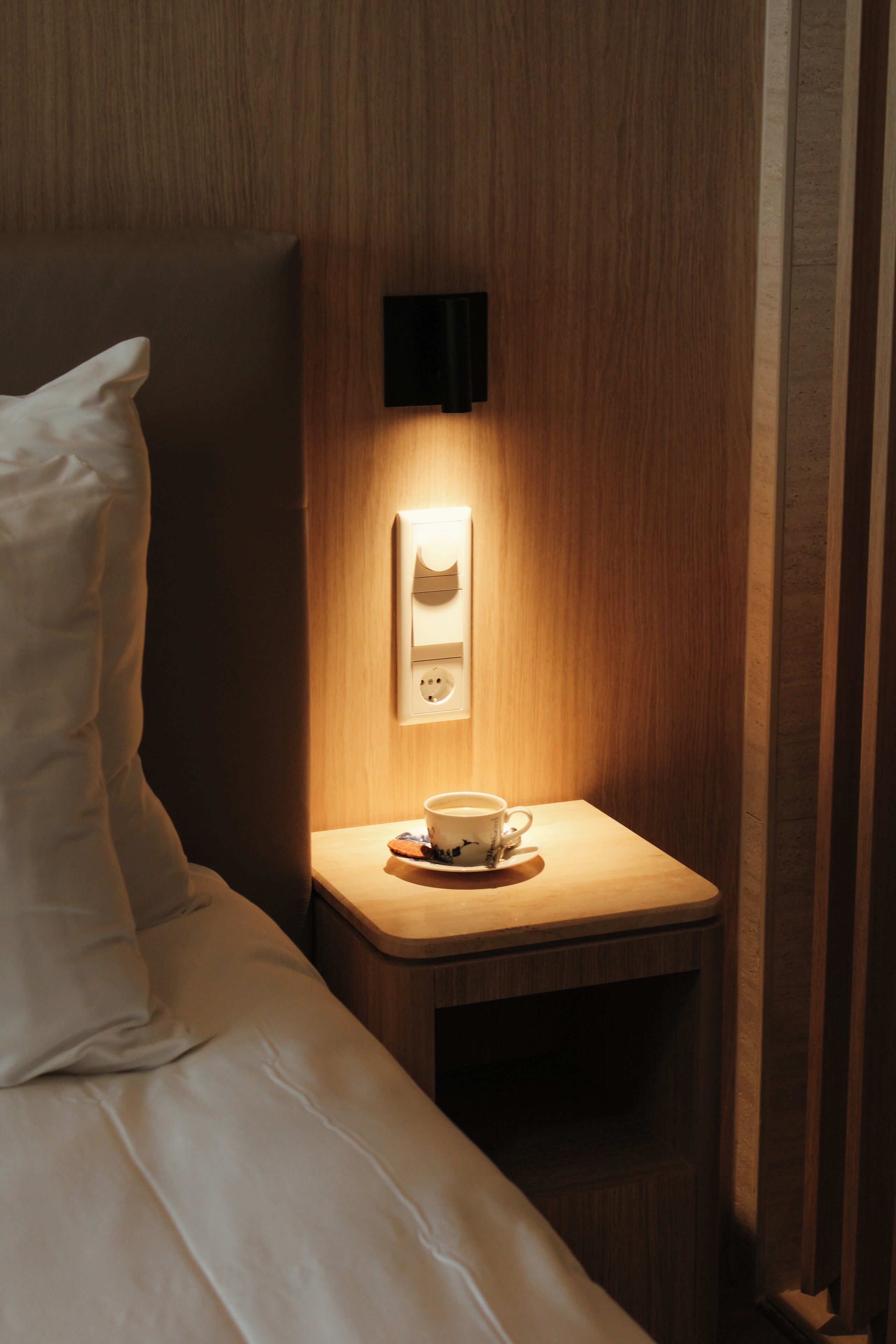 Een hotelkamer met een net opgemaakt bed, een nachtkastje met een kop koffie, en een wandlamp.
