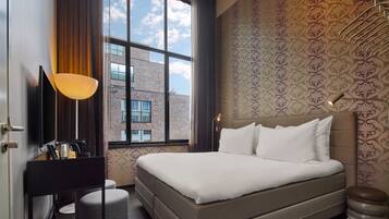 A modern hotel room with a large bed, a TV, a desk with a lamp, and a view of the sky through the window.