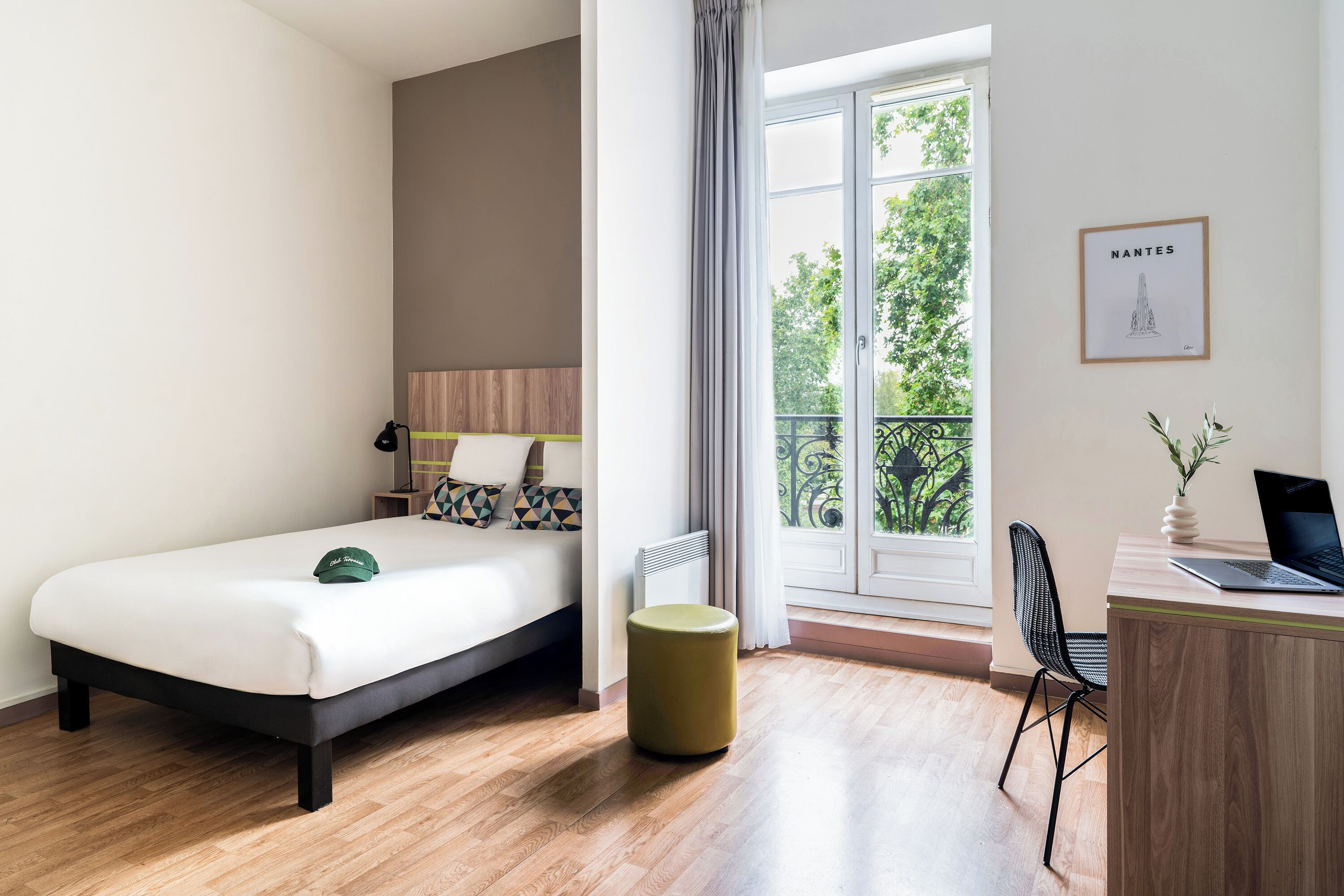 A hotel room with a bed, a desk with a laptop, a chair, a radiator, and a window with a view of trees.