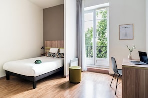Habitación de hotel con una cama, un escritorio con una computadora portátil, una silla, un radiador y una ventana con vista a árboles.