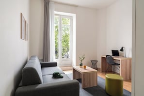 Un salon moderne avec un canapé gris, une table basse en bois, un petit bureau avec un ordinateur portable, une chaise et un pouf jaune.