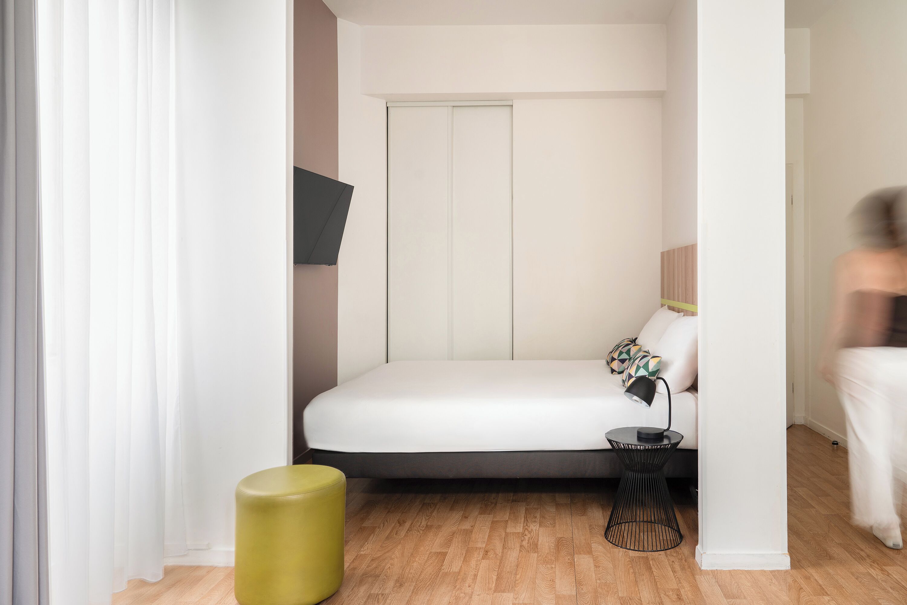 A bedroom with a bed, a bedside table, a lamp, a small stool, and a wardrobe.
