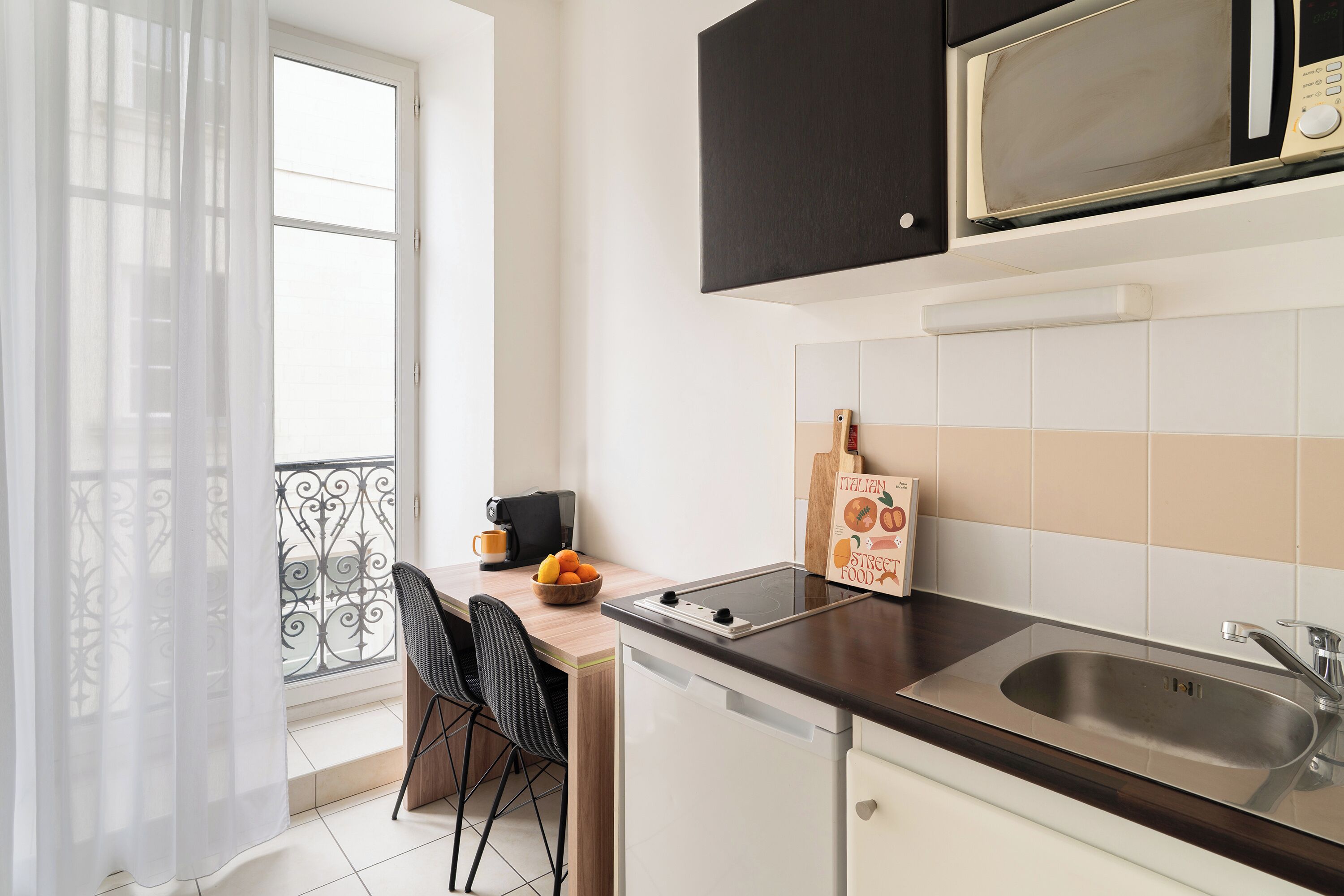 Une cuisine compacte équipée d’un four à micro-ondes, d’un évier et d’un petit coin repas avec une chaise et un saladier de fruits.