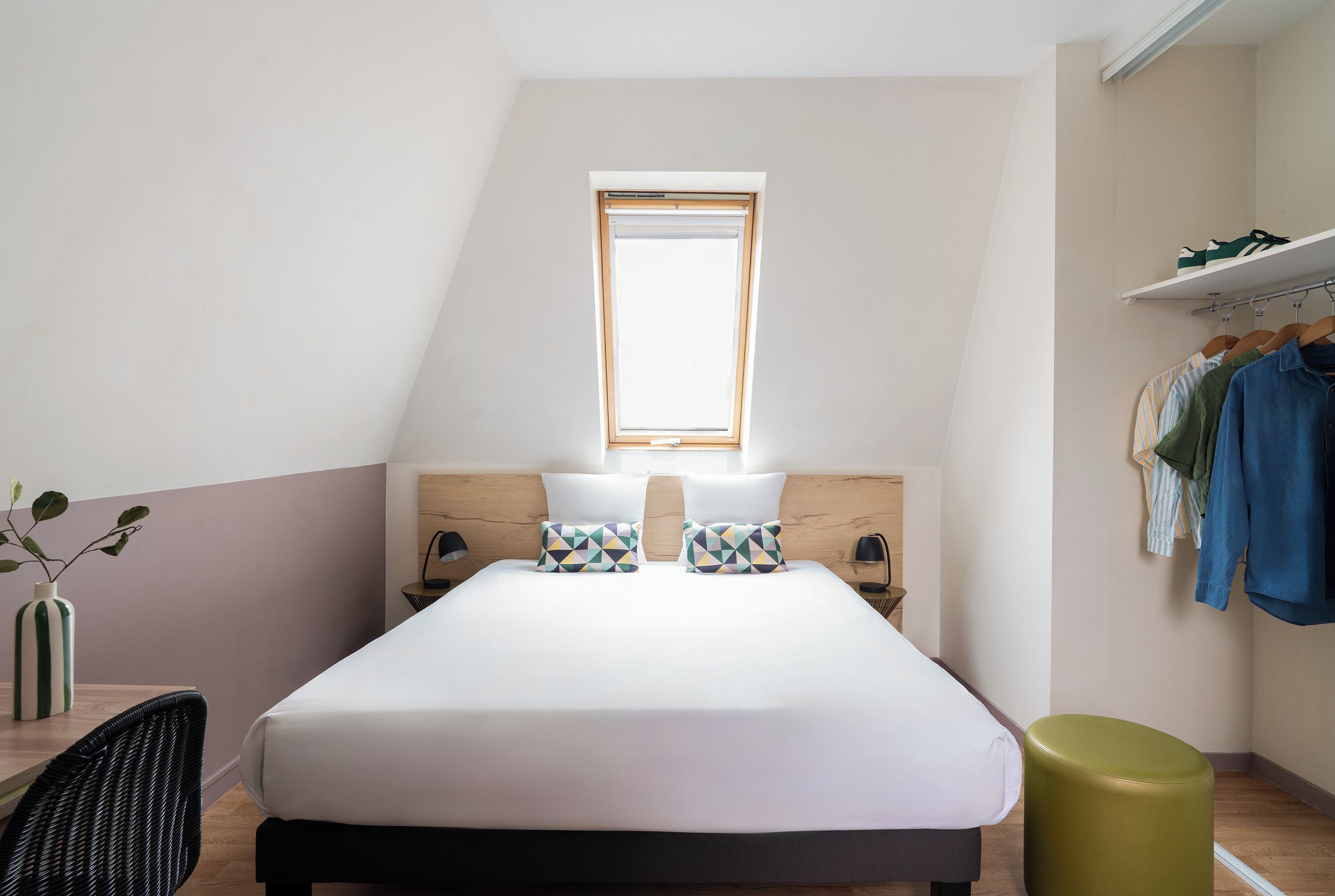 A bedroom with a bed, two pillows, a bedside table, a lamp, a small plant, a chair, a stool, a window, and a clothing rack.