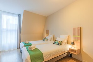 Une chambre d’hôtel avec deux lits, une table de chevet avec une lampe et une grande fenêtre avec des rideaux légers.