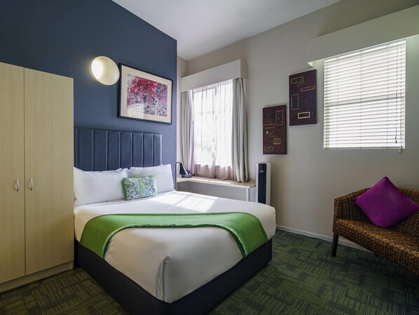 Habitación de hotel con cama, armario, ventana con cortinas, una silla y obras de arte en las paredes.