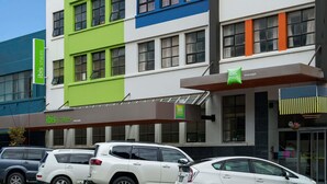 Un hotel de varios pisos con fachada moderna, que presenta una combinación de colores verde, azul y naranja. Hay varios coches estacionados frente al hotel, y una señal indica que forma parte de la cadena de hoteles ibis Styles.