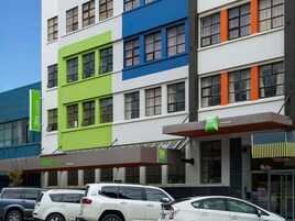 Un hotel de varios pisos con fachada moderna, que presenta una combinación de colores verde, azul y naranja. Hay varios coches estacionados frente al hotel, y una señal indica que forma parte de la cadena de hoteles ibis Styles.