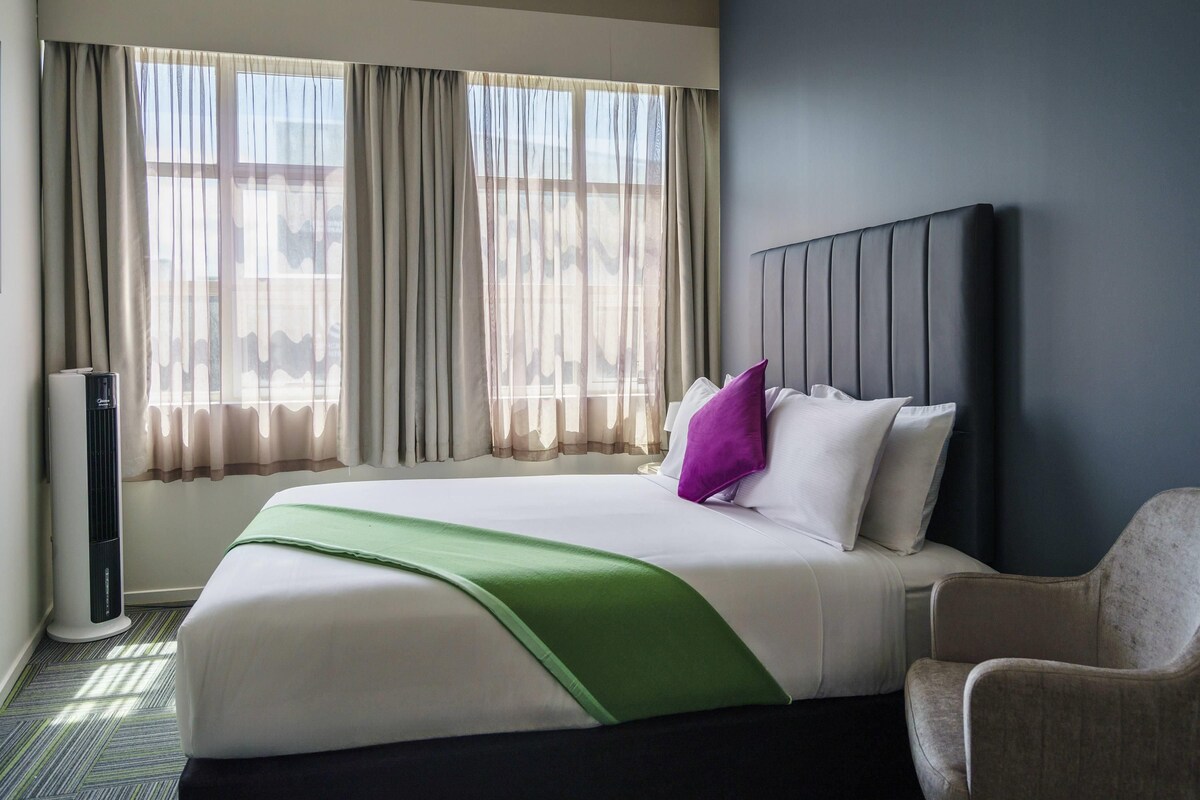 Una habitación de hotel con cama, una silla, ventana con cortinas y una unidad de pared.