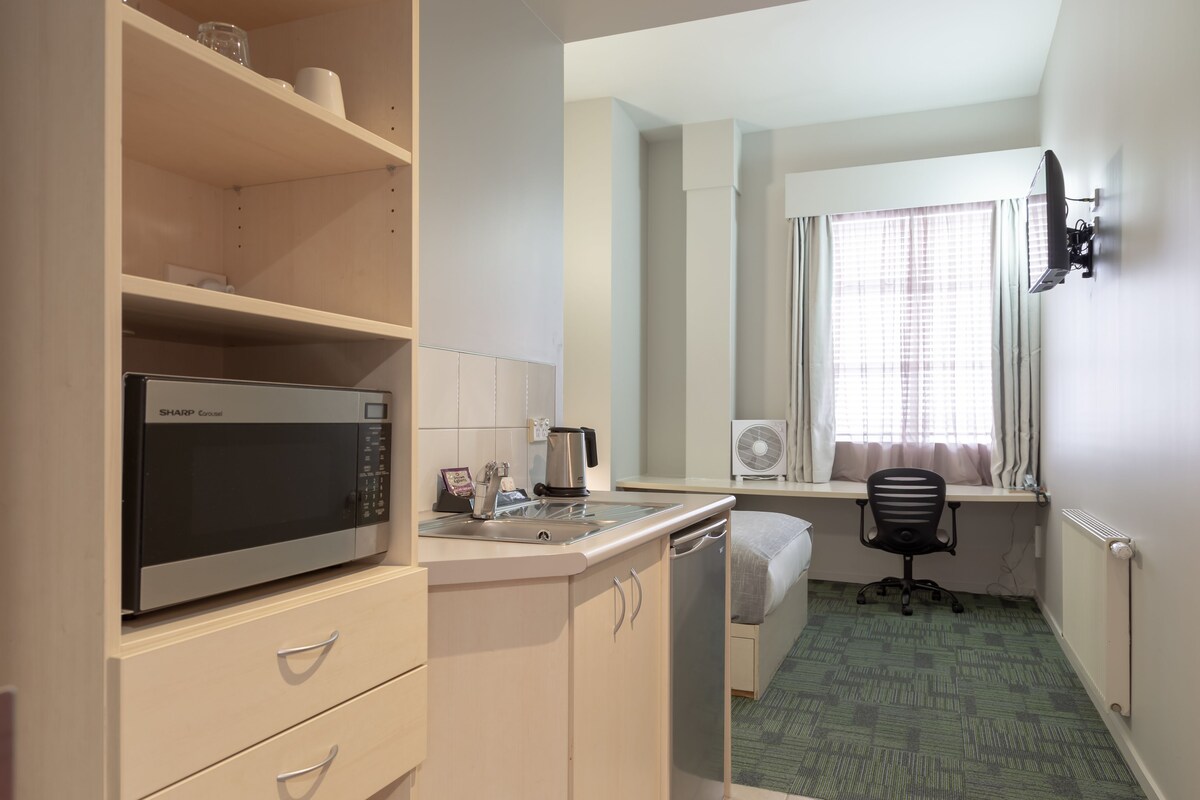 Habitación de hotel compacta con kitchenette, una cama individual, un escritorio y ventana con cortinas.