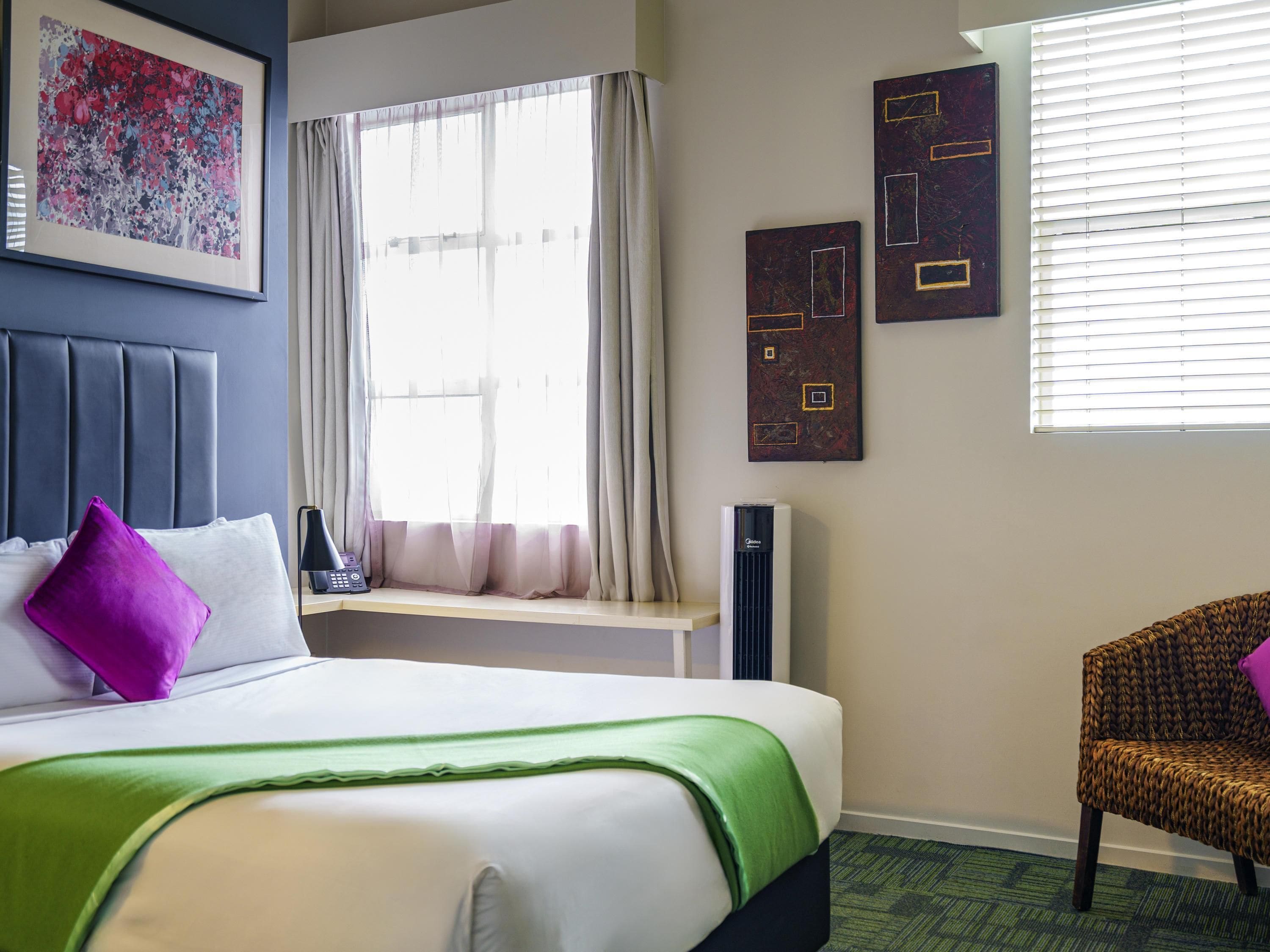 Una habitación de hotel con una cama, una ventana con cortinas, una silla y obras de arte en las paredes.