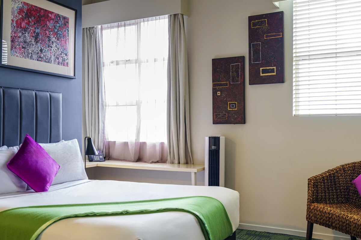 Una habitación de hotel con una cama, una ventana con cortinas, una silla y obras de arte en las paredes.