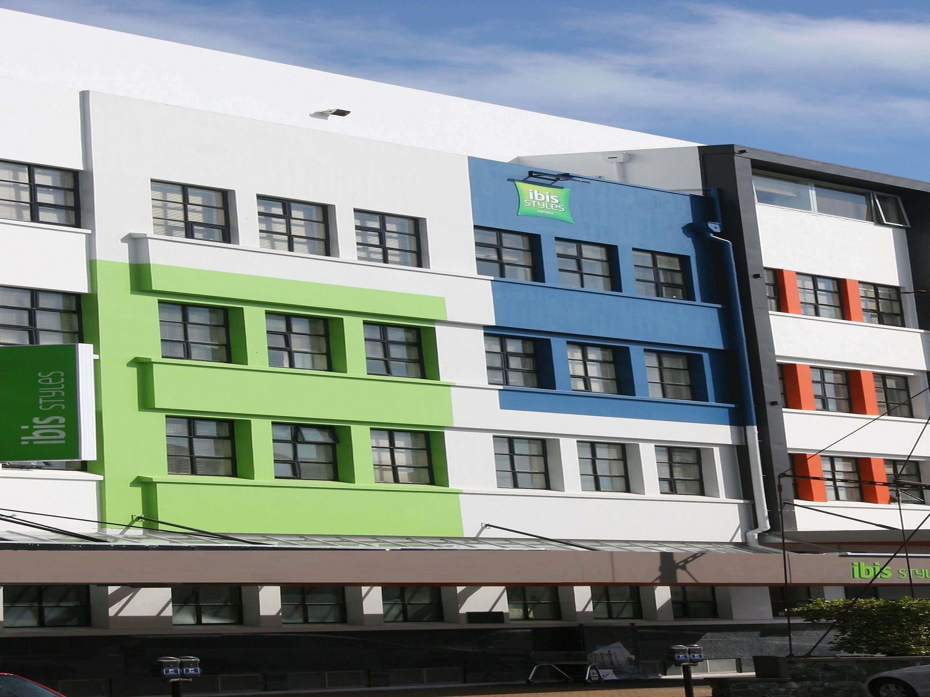A multi-story hotel with a modern design, featuring a green and blue facade, multiple windows, and a sign indicating it is an ibis hotel.