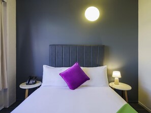 Una habitación de hotel con una cama, teléfono en la mesita de noche, lámpara y luz de pared redonda.