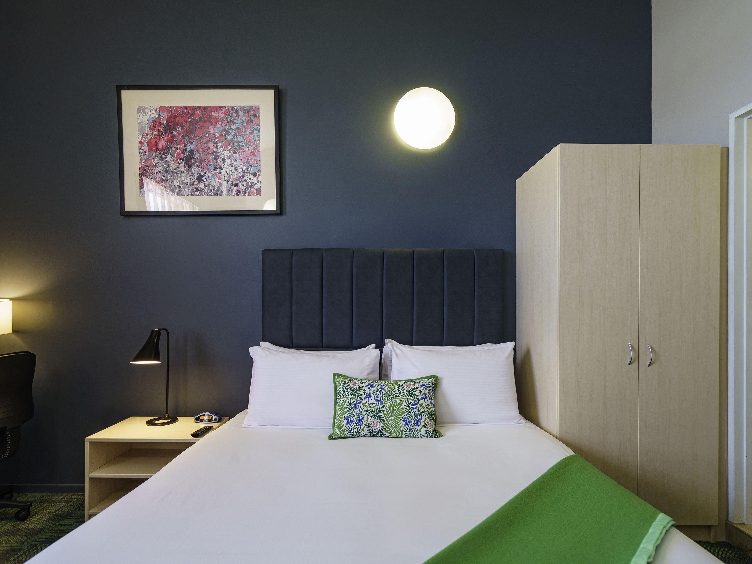 A hotel room with a bed, a nightstand, a lamp, a wardrobe, and a framed picture on the wall.